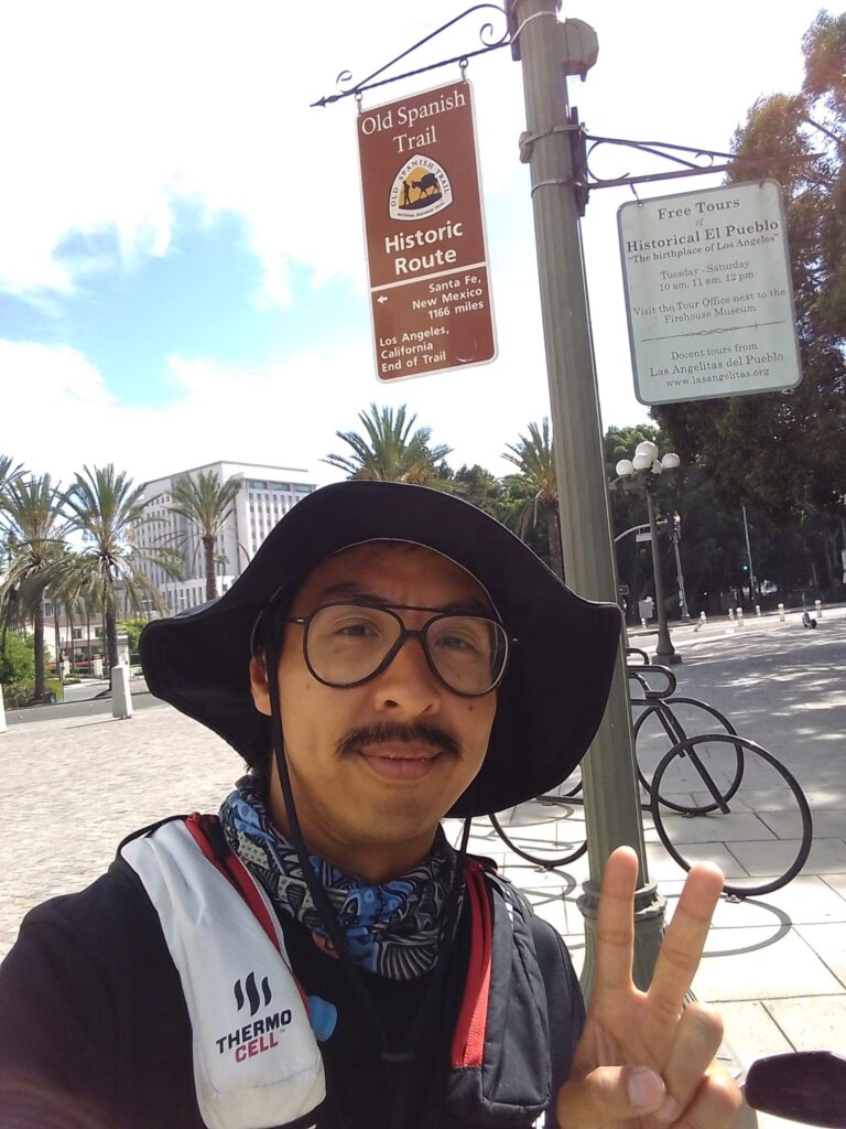 the author giving a peace sign, background sign "Old Spanish Trail Historic Route, Los Angeles, California, End of trail"